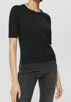 Kobieta ubrana w czarny dzianinowy top z krótkim rękawem i bocznymi rozcięciami oraz ciemnoszare jeansy, uzupełnione złotym naszyjnikiem z łańcuszka.