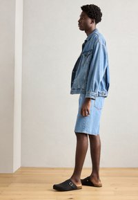 Jeans jakna i šortsevi svijetloplave nijanse. Crni klompi s detaljem kopče. Pozadina od prirodnog drvenog poda. Jednostavan, suvremeni dizajn.