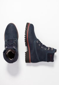 Blå mocka ank stövlar med svarta gummisålar, texturerad finish, metallöglor och en röd accent runt sålen. Fodrad innersula synlig.