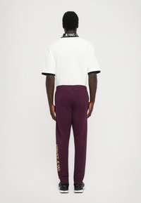 O conjunto masculino apresenta uma polo branca de manga curta com detalhes pretos e calças de moletom roxas. As calças têm "Versace Jeans" impresso em dourado.