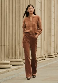 Jachetă zip-up fuzzy maro, pantaloni evazați maro și pantofi cu toc strălucitori. Modelul stă în fața unui fundal cu coloane, etalând ținuta.