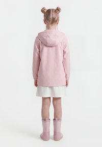 Kind met blond haar in dubbele knotjes, gekleed in een roze gevoerde jas, witte rok en roze regenlaarzen, staande met de rug naar de kijker op een witte achtergrond.