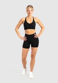 Reggiseno sportivo nero con design arricciato, abbinato a pantaloni corti neri a vita alta. Entrambi i capi sono realizzati in tessuto elasticizzato e morbido. Sneakers bianche.