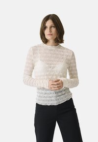 Langärmlige, transparente Bluse in hellem Creme mit strukturierten Blumenmustern und Rüschenmanschetten. Kombiniert mit schwarzen Hosen.