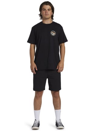 T-shirt nera in cotone con un logo rotondo sul petto, abbinata a pantaloni corti neri. Calzini bianchi a girocollo e scarpe nere con accenti bianchi.