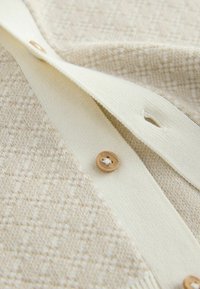 Cream knit cardigan featuring a textured pattern, a smooth cream band, and a brown button detail. Focus on soft fabric and geometric design.