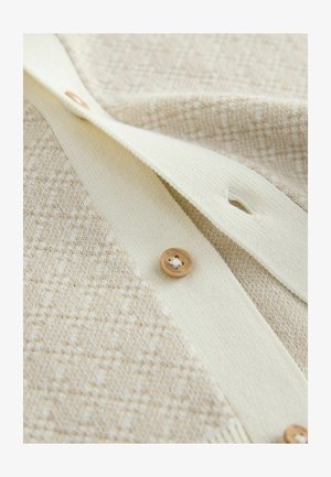 Krēmkrāsas adīts kardigans ar teksturētu rakstu, gludu krēmkrāsas joslu un brūnu pogu detaļu. Uzsvars uz mīkstu audumu un ģeometrisku dizainu.