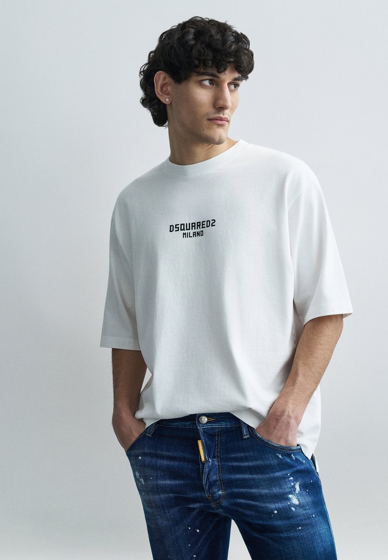 T-shirt bianca oversize con maniche corte e logo nero "DSQUARED2 MILANO"; abbinata a jeans denim blu strappati con schizzi di vernice.
