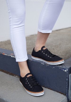 Persona que lleva zapatillas negras y marrones con cordones y pantalones blancos, pisando un bordillo de hormigón azul al aire libre.