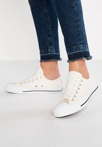 Baskets en toile écrues avec une finition texturée, présentant des œillets argentés, des bouts blancs et des bandes d'accent noir sur la semelle.