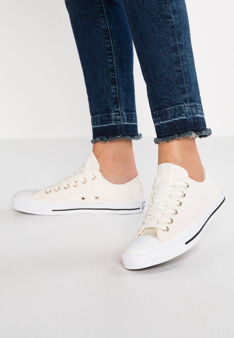 Baskets en toile écrues avec une finition texturée, présentant des œillets argentés, des bouts blancs et des bandes d'accent noir sur la semelle.