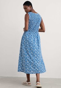 Maxi dress featuring a blue and white leaf pattern, sleeveless design, smocked bodice, and flowing skirt, worn with light-colored sandals.