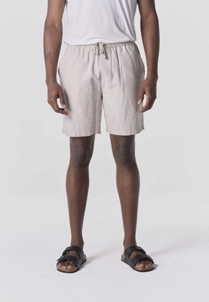 Osoba ubrana w jasnobeżowe szorty ze ściągaczem, biały t-shirt i czarne sandały, stojąca na tle jednolitego szarego tła.