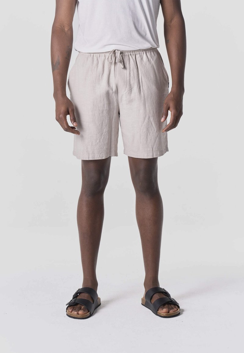 Isik, kes kannab helebeeži värvi paelaga lühikesi pükse, valget t-särki ja musti sandaale, seisab ühevärvilise halli tausta ees.