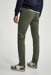 Uomo in pantaloni verde oliva, felpa blu navy e sneakers beige-marroni che si trova su uno sfondo chiaro e uniforme.