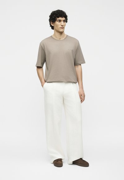 Νεαρός άνδρας με σγουρά μαλλιά, φοράει μπεζ κοντομάνικο πουκάμισο, λευκό παντελόνι με φαρδιά μπατζάκια και καφέ παπούτσια, στέκεται με τα χέρια στις τσέπες.