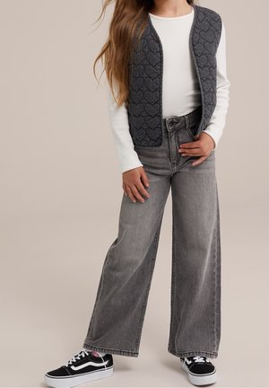 Dziecko w białej koszuli z długim rękawem, ciemnoszarej kamizelce w serduszka, szerokich szarych dżinsach i czarno-białych trampkach.