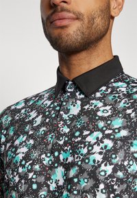 Svart blomstrete polo-skjorte med et utvalg av turkise og hvite blomsterdesign, med en kontrasterende svart krage og knappdetaljer.