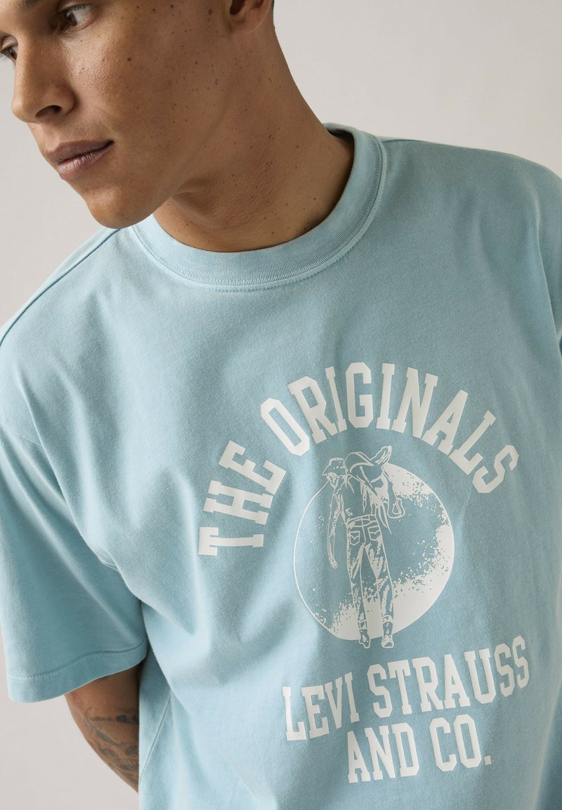 Hombre con piel marrón clara y tatuaje que lleva una camiseta azul claro de Levi Strauss con "The Originals" y un gráfico de un hombre cargando pantalones vaqueros.