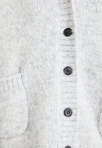 Cardigan lavorato a maglia di un grigio chiaro, con un tessuto testurizzato, chiusura a bottoni e una tasca a toppa sul davanti. Bottoni neri creano un contrasto.