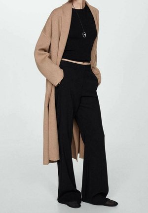 Persoon die een lange camelkleurige jas, een zwart cropped top, wijde zwarte broek, zwarte schoenen en een lange ketting met hanger draagt, staand met handen in de zakken.