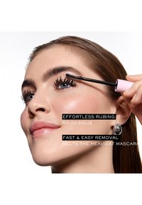 Femme appliquant du mascara sur les cils supérieurs, mettant en valeur une peau lisse, un maquillage naturel et une boucle d'oreille argentée en forme de créole, avec un texte superposé indiquant les bénéfices du produit.