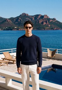 Homme portant des lunettes de soleil, un pull bleu marine et un pantalon blanc, debout près d’une piscine avec l’océan et des collines rocheuses en arrière-plan par une journée ensoleillée.