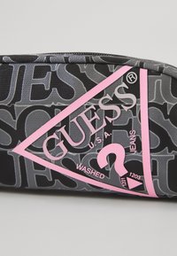 Svart påse med ett upprepande mönster av "GUESS"-logotypen. Den har en triangulär rosa logotyp med "WASHED" och en frågetecken-design.
