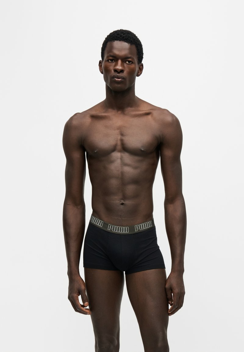 Homme noir athlétique debout, portant un boxer Puma noir avec une ceinture élastique dorée, sur un fond gris clair uni.