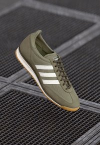 Olive-green athletic shoe with white stripes, mesh material, and gum rubber sole. Features a rounded toe and lace-up front.