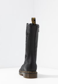 Vysoká černá kožená bota s květinovou výšivkou, žlutým švem, silnou průhlednou podrážkou a černým poutkem s žlutým textem.