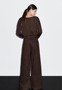 Bruine geribbelde trui en wijde bruine broek, met een hoge taille en achterzak, gezien vanaf de achterkant tegen een lichte achtergrond.