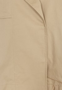 Tissu beige avec coutures et surpiqûres visibles, montrant des détails en gros plan de la fabrication et de la texture du vêtement.