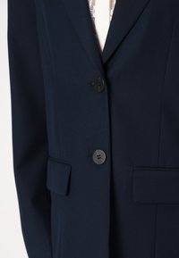 Marineblazer laget av teksturert stoff, med tre sorte knapper, to frontlommer og en skreddertilpasset passform. Subtile sømmer synlige.