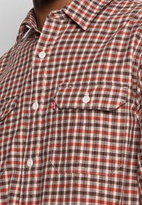 Camicia a quadri marrone, arancione e crema con bottoni bianchi e taschino sul petto con bottone, indossata da una persona.