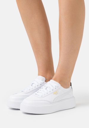 Vita sneakers med plattformssula, slät läderöverdela, guldlogotypdetalj och svart accent vid hälen. Snörning går genom öglor.