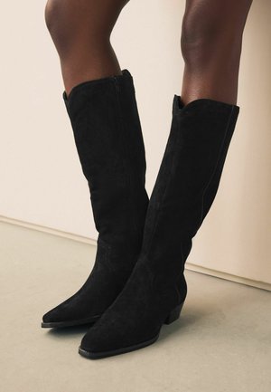 Botas altas de ante negro hasta la rodilla con punta afilada y tacón bajo tipo bloque. Tienen cremallera lateral y un sutil detalle de costura a lo largo del caño.