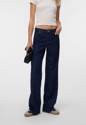 Person som har på seg mørkeblå vide jeans og hvit cropped t-skjorte, holder en svart clutchveske, med joggesko på grå bakgrunn.
