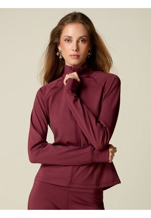 Frau mit langen Haaren, die eine bordeauxrote Jacke mit Reißverschluss und passende Hose trägt, blickt nach vorne, eine Hand nahe am Kinn und bunte Ohrringe sichtbar.