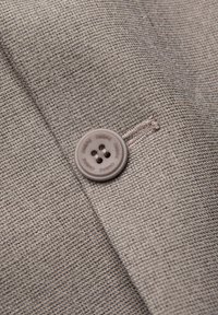 Tissu gris à tissage fin, featuring un bouton rond gris clair à quatre trous, attaché à une poche avec un détail de couture subtil.