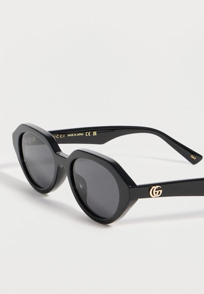 Lunettes de soleil Gucci noires avec montures octogonales arrondies, verres foncés et logo double-G doré sur les branches.