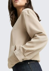 Blouse beige à manches longues avec une coupe décontractée, présentant des détails cousus, une poche latérale et des accents de boutons sur le poignet. Associée à un jean foncé.