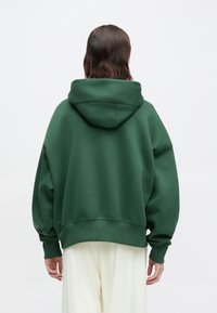 Person med skulderlangt mørkt hår iført en oversized mørkegrønn hettegenser og vide hvite bukser, stående vendt bort fra kameraet mot en ensfarget bakgrunn.