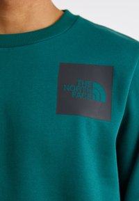Teal genser med ribbestrikket hals og en svart rektangulær lapp med "The North Face"-logo i teal bokstaver.
