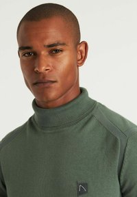 Olivgrüner Rollkragenpullover aus geripptem Stoff, mit einer taillierten Passform und einem kleinen Logopatch auf der linken Brust.