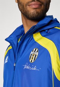 Blauw-gele ritsjas met Juventus-logo, een gladde textuur, rits aan de voorkant en een kenmerkend detail op de borst.