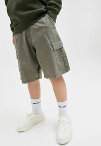 Kargo kratke hlače iz olivno zelene tkanine, z bočnimi žepi in sproščenim krojem, v kombinaciji z belimi supergami in belimi nogavicami.