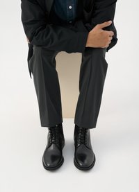 Bocanci negri din piele cu vârfuri rotunde, sistem de închidere cu șireturi și textură netedă, purtați cu pantaloni întunecați și o jachetă neagră.