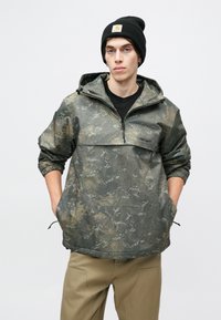 Kamuflážová mikina s kapucňou, vodotesný materiál, šedé a zelené odtiene, nastaviteľná kapucňa, predná vrecko, elastické manžety a lem. Skombinované s béžovými nohavicami.