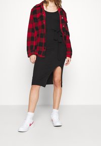 Robe noire côtelée avec une fente latérale, cintrée à la taille, portée sous une chemise-veston à carreaux rouges et noirs. Portée avec des baskets blanches.